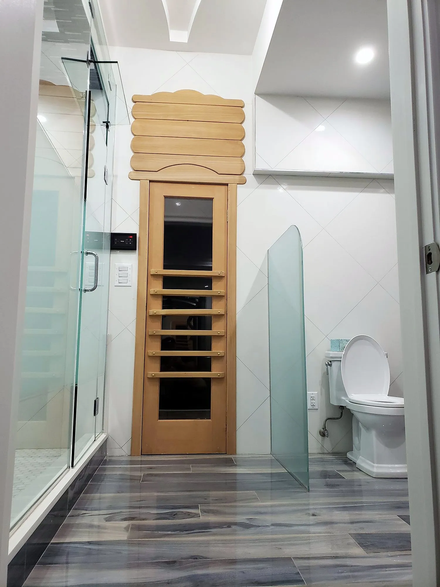 Finished bathroom with blue-grey wood-look porcelain floor, glass partitions and a cedar sauna door