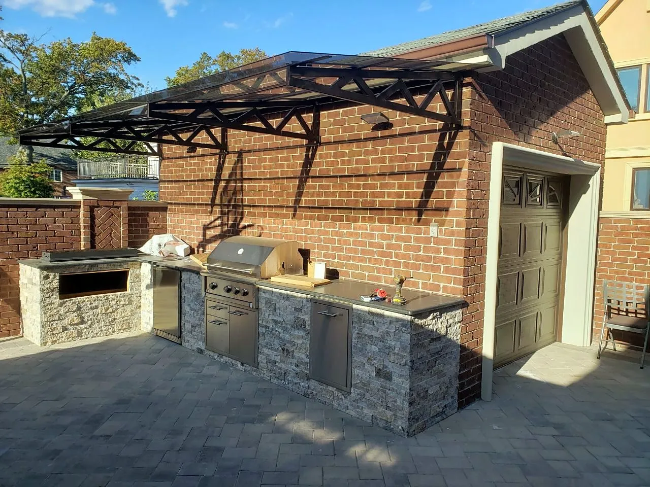 Brick garage with a steel-framed glass pavilion roof and a stone outdoor kitchen in a Queens backyard